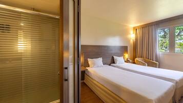 Habitación de hotel con dos camas, cabecera de madera, mesita de noche con lámpara y ventana con cortinas.
