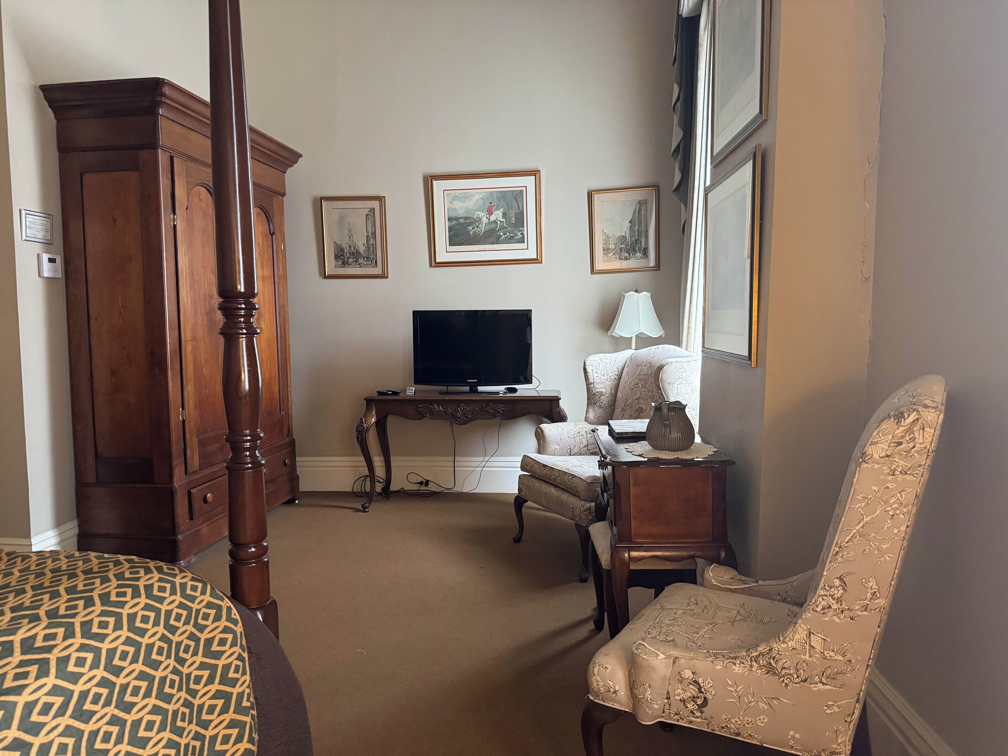 A room with a bed, a wooden wardrobe, a television on a stand, a desk with a lamp, and three framed pictures on the wall.