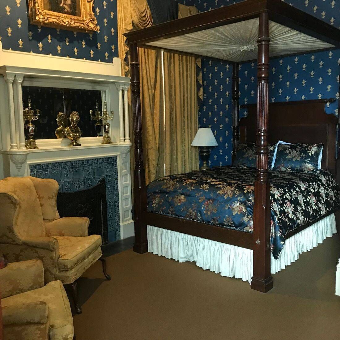 A four-poster bed with a floral bedspread, a fireplace with decorative items, and a classic armchair.