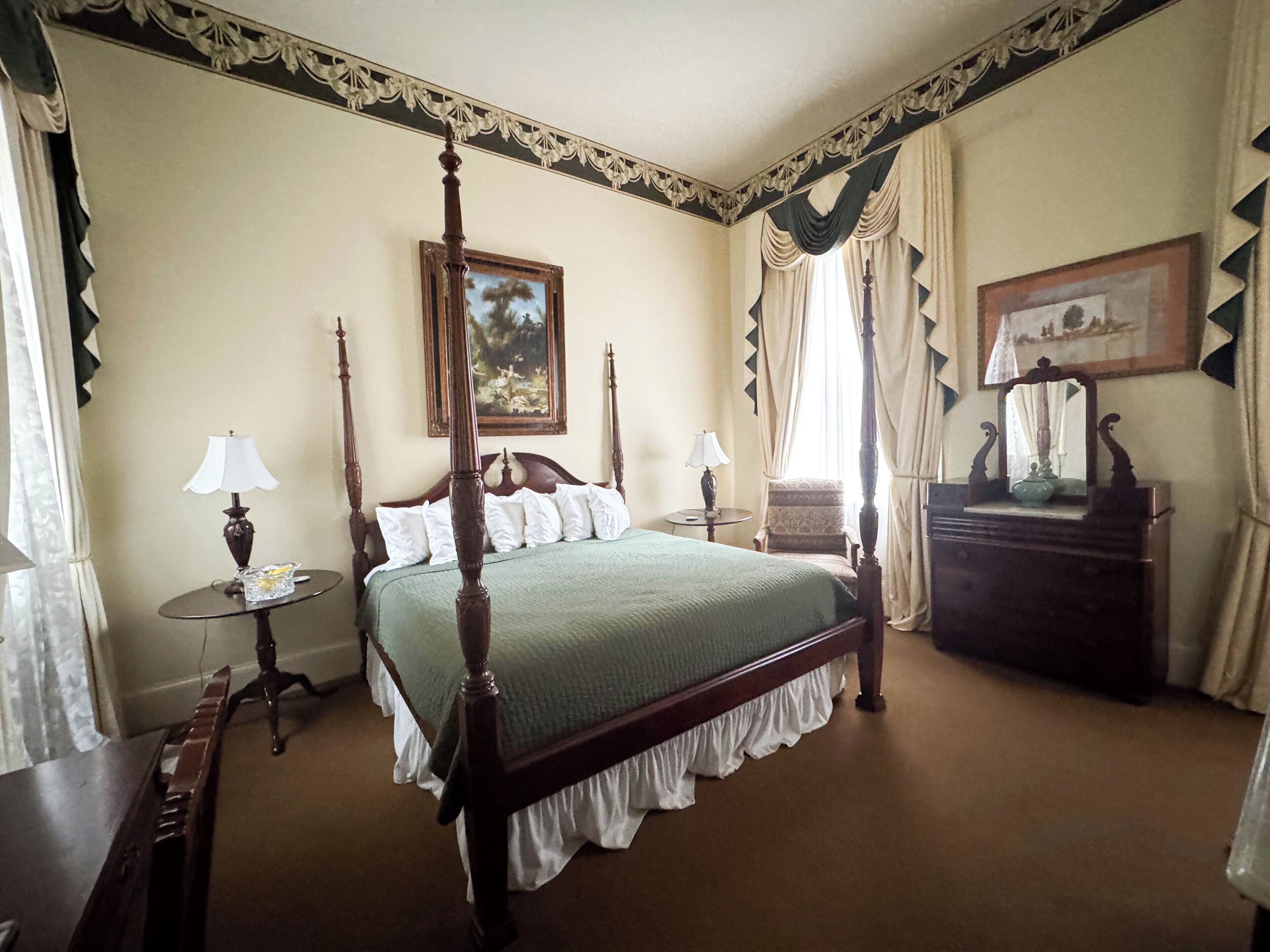 A four-poster bed with a green bedspread, two bedside tables with lamps, a chair, a dresser, and a painting on the wall.
