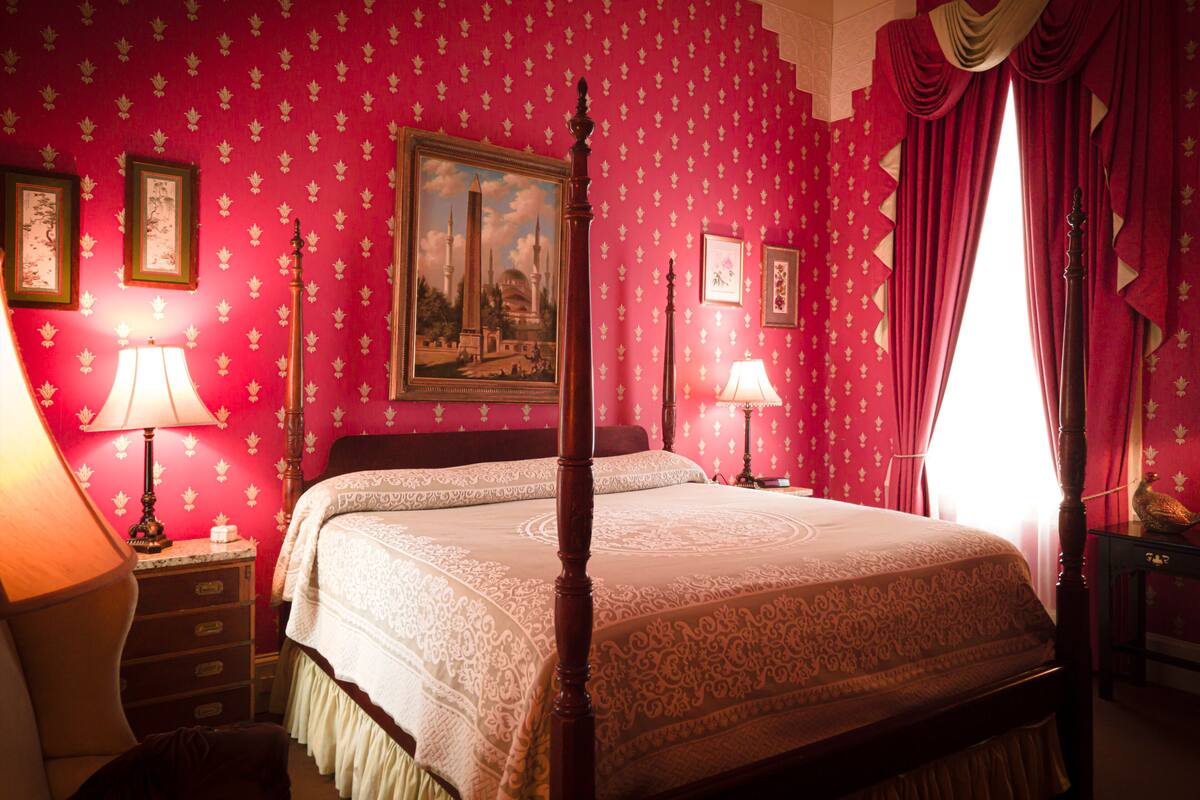 A bedroom with a four-poster bed, red patterned wallpaper, and a large window with red curtains.