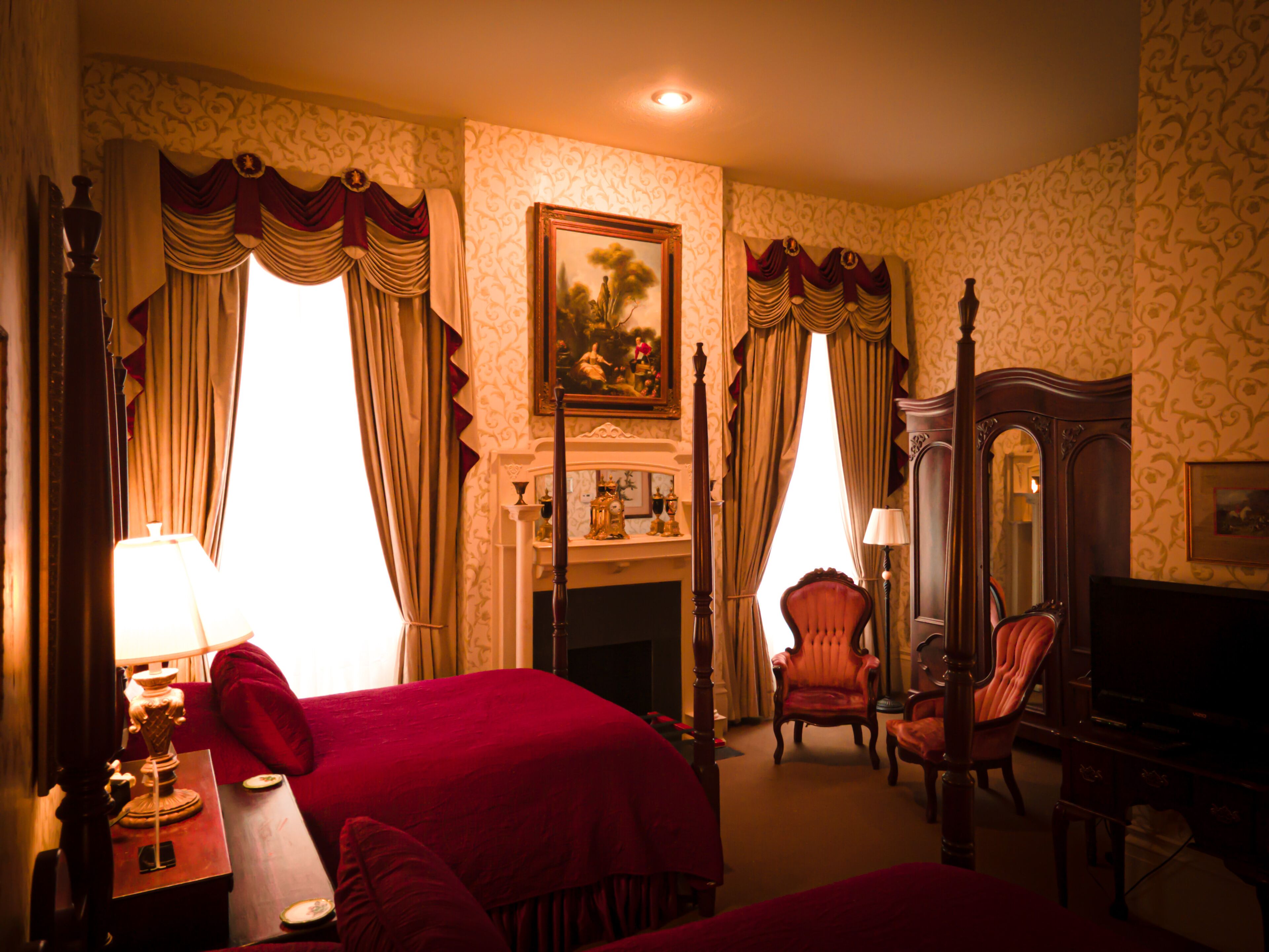 A hotel room with a fireplace, two red upholstered chairs, a wooden bed, and a large mirror.