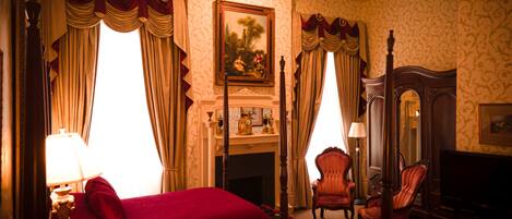 A hotel room with a fireplace, two red upholstered chairs, a wooden bed, and a large mirror.