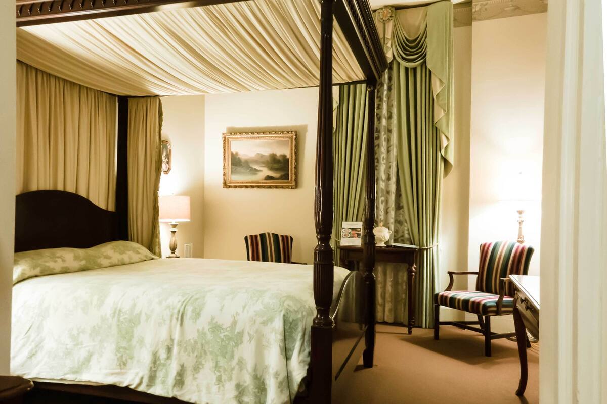 A four-poster bed with a canopy, a bedside table with a lamp, a chair, and a small table with a vase.