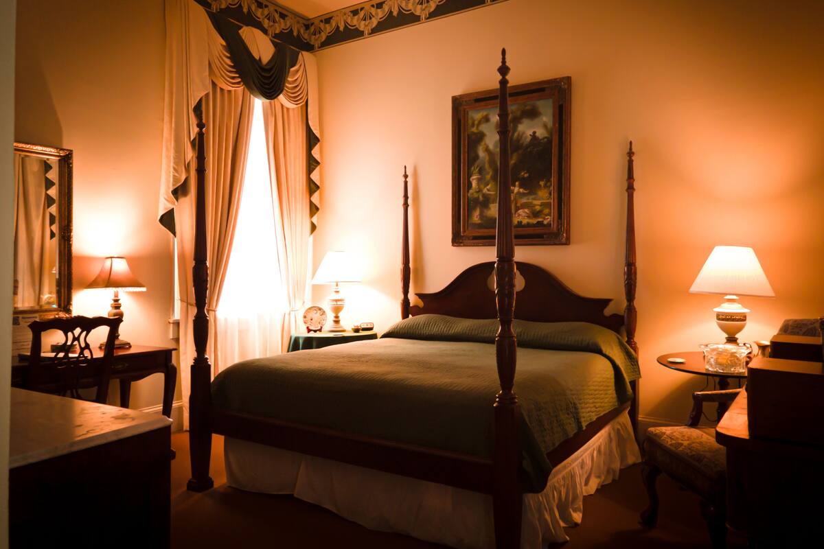 A four-poster bed with a green bedspread, two bedside lamps, a nightstand with a clock, a chair, and a painting on the wall.