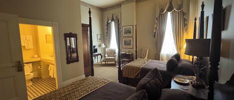 Une chambre d’hôtel avec deux lits, un coin salon comprenant une chaise et une table, une salle de bain visible à travers une porte ouverte, et un grand miroir.