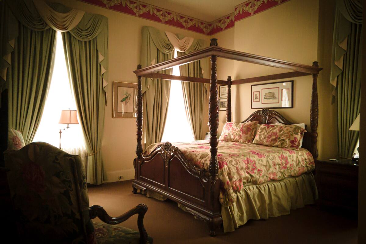A four-poster bed with floral bedding, a chair with a floral pattern, and a lamp on a bedside table.