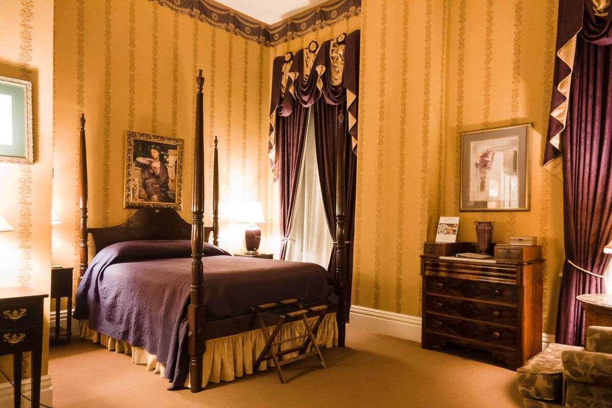 A four-poster bed with a canopy, a dresser, a chair, and a desk in a room with striped wallpaper.