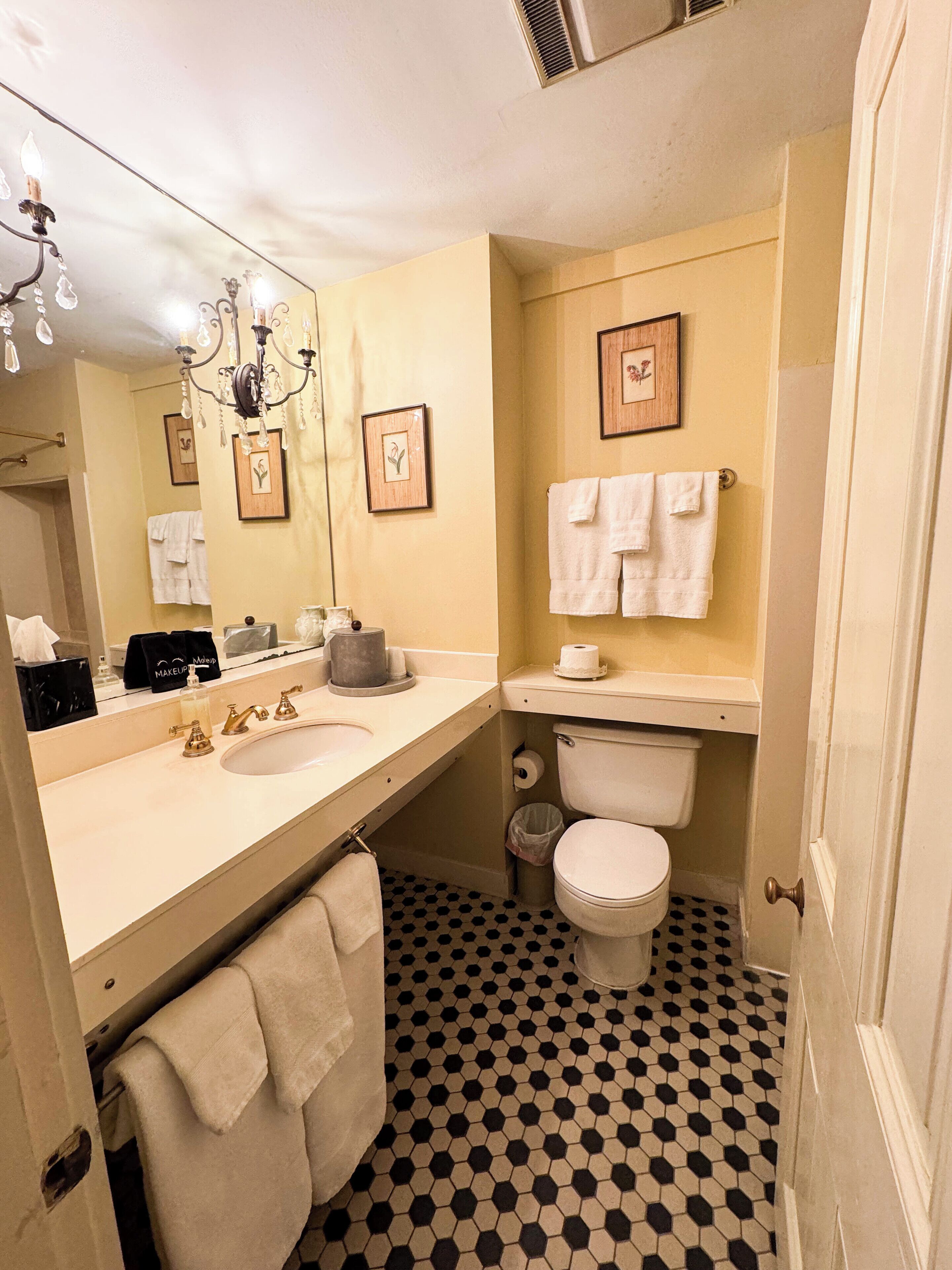 Une salle de bain avec un lavabo, des toilettes et un miroir. Des serviettes sont accrochées au mur et un lustre se trouve au-dessus du lavabo.