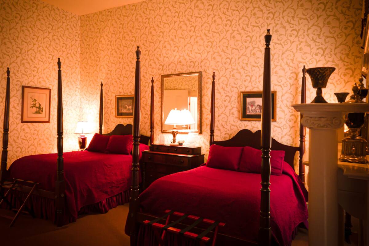 A four-poster bed with a red bedspread, a nightstand with a lamp, and a mirror on the wall.