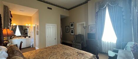 Habitación de hotel con una cama grande, un escritorio, una silla, un tocador con espejo y una ventana con cortinas.