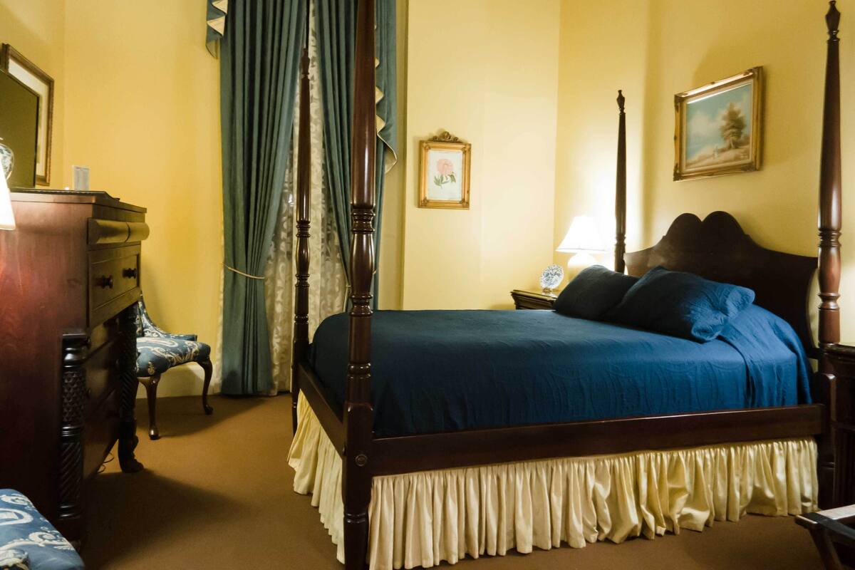 A four-poster bed with a dark wooden frame and a blue bedspread, a nightstand with a lamp, a dresser, and a chair with a patterned cushion.