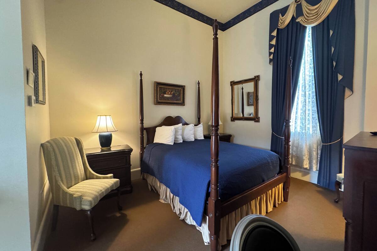 A four-poster bed with a blue bedspread, a chair, a nightstand with a lamp, and a mirror on the wall.