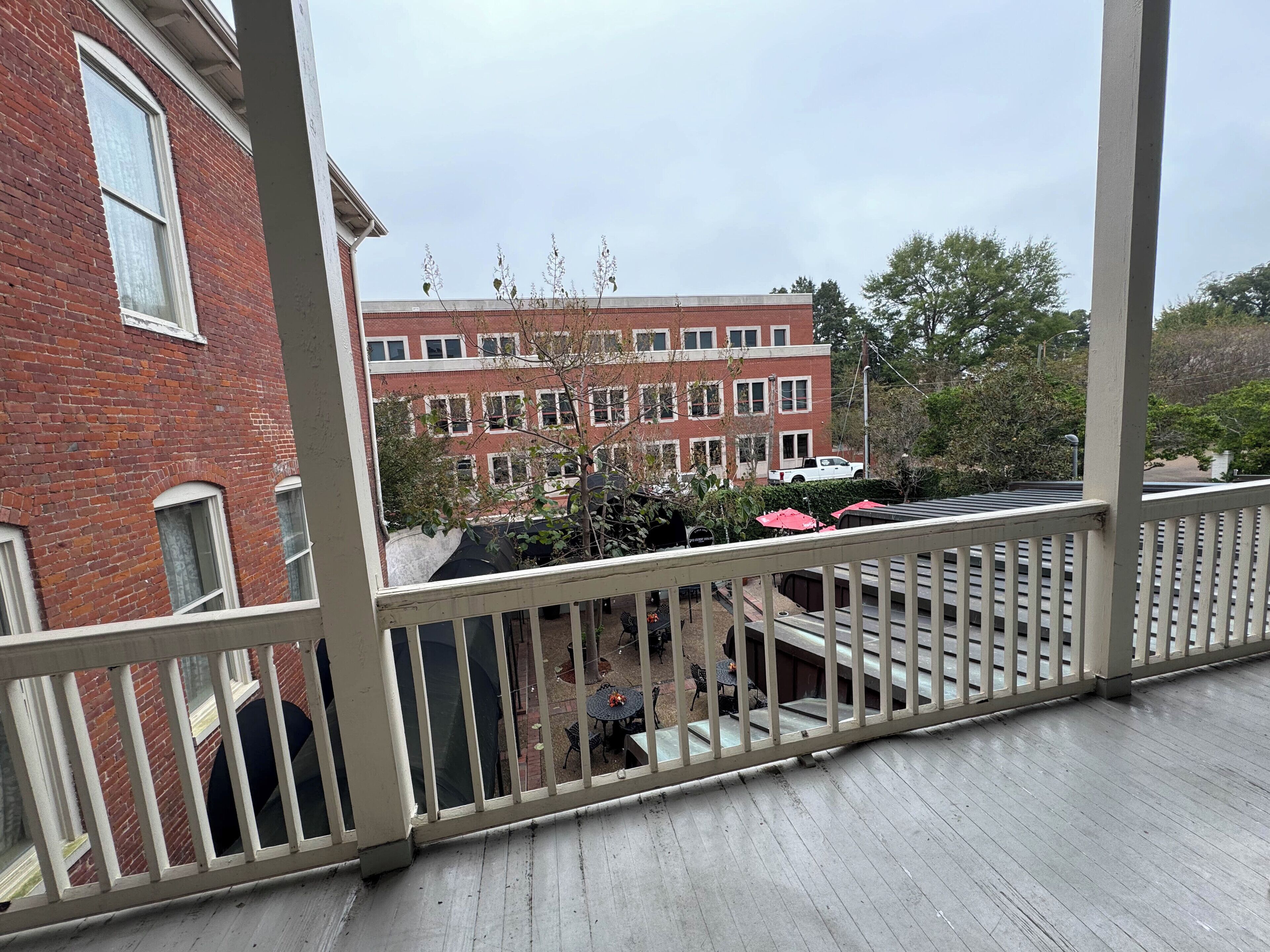 Vista da un balcone su un edificio in mattoni con numerose finestre, un’area patio con tavoli e sedie, e un’auto bianca parcheggiata sullo sfondo.