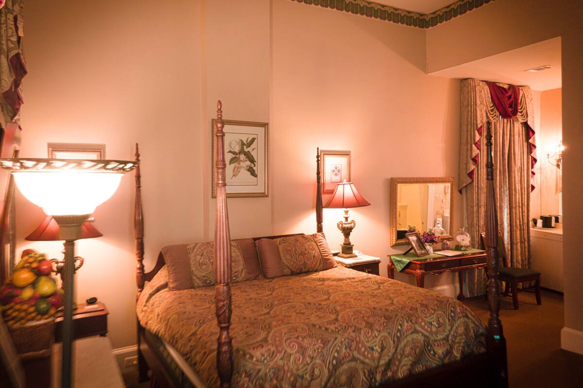 A traditional bedroom with a four-poster bed, two bedside lamps, a mirror, and a side table with a lamp.