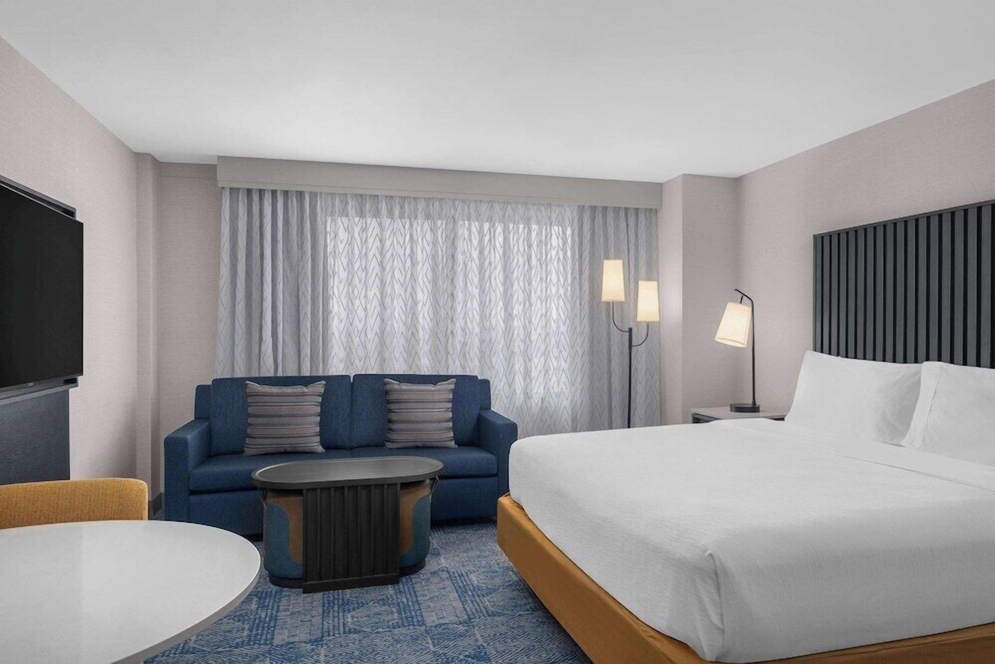 A hotel room with a bed, a sofa, a round coffee table, a TV, and a window with curtains.