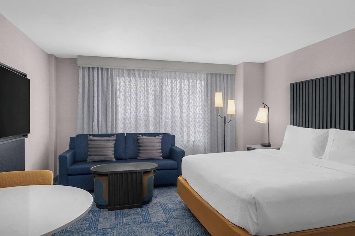 Quarto de hotel com cama, sofá, mesa de centro redonda, televisão e janela com cortinas.