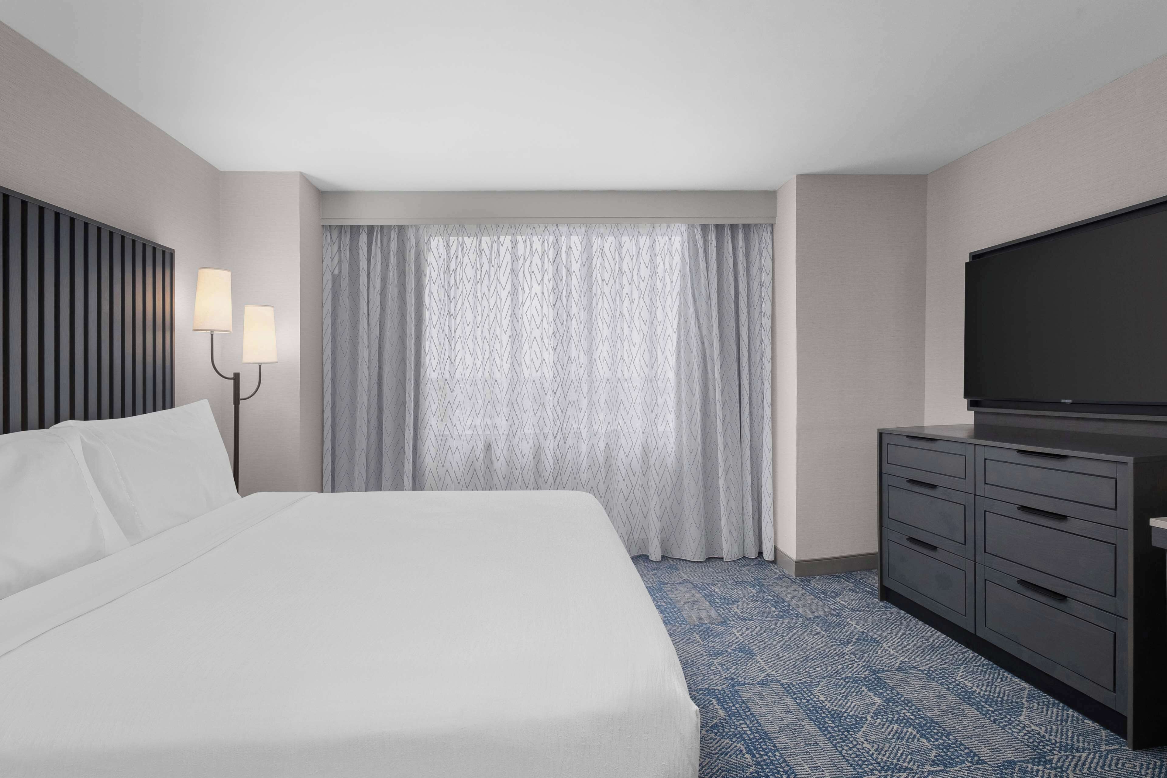 Una habitación de hotel con una cama grande, un televisor de pantalla plana, un aparador y una ventana con cortinas.