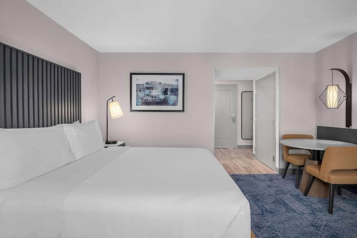 Quarto de hotel com cama, mesa de cabeceira, luminária, quadro emoldurado e um espaço de refeições com cadeiras.