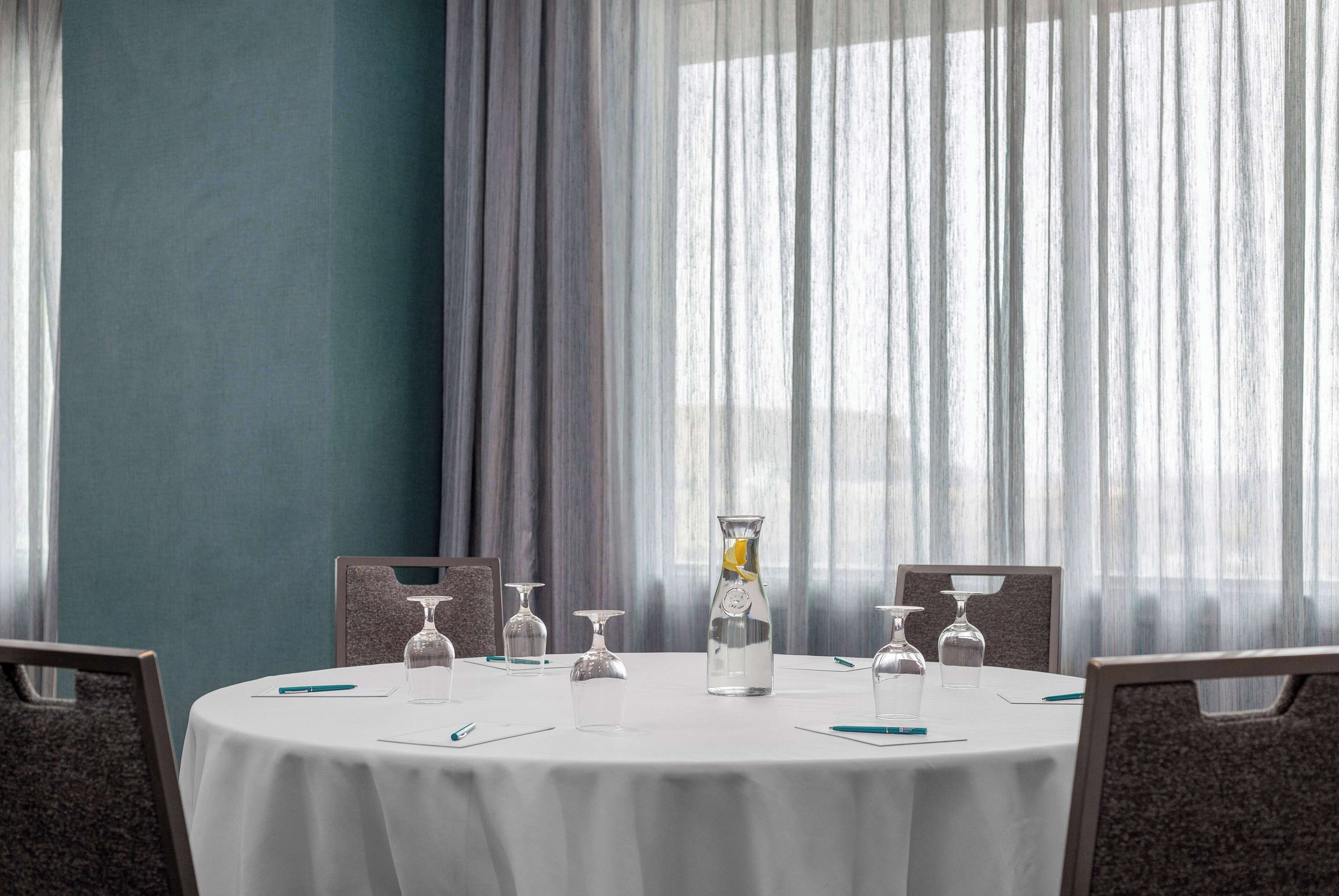 Een ronde tafel met een witte tafelkleed, omringd door stoelen, voor een raam met dunne gordijnen.