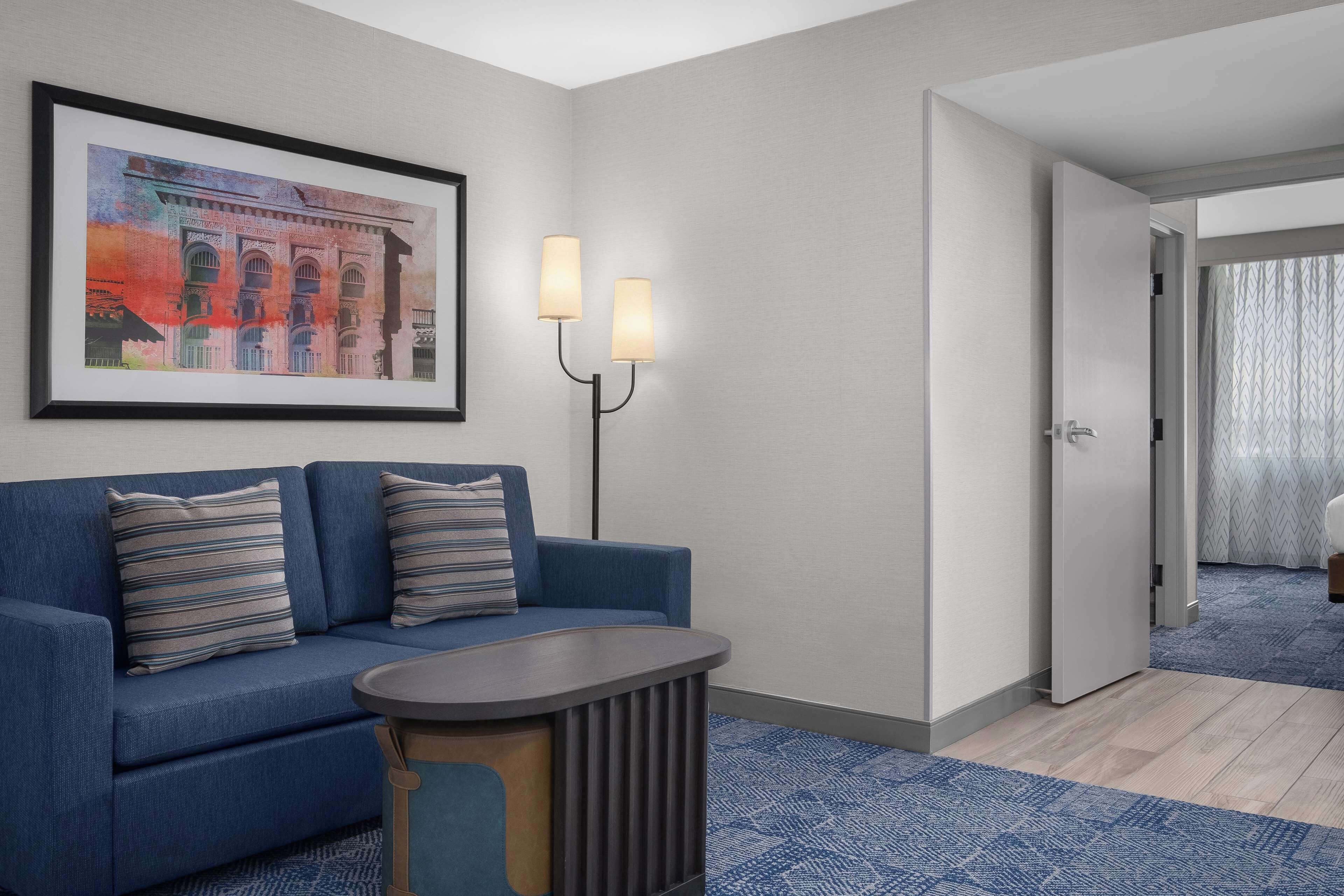 A hotel room with a blue sofa, a framed picture on the wall, a standing lamp, and a door leading to another room.