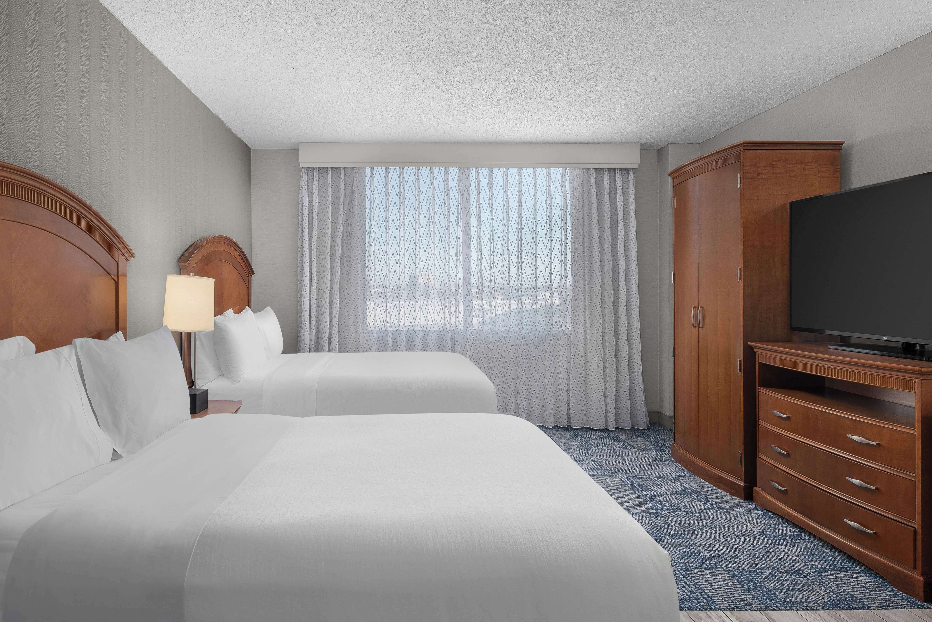Una habitación de hotel con dos camas, un televisor, un armario de madera y una ventana con cortinas translúcidas.