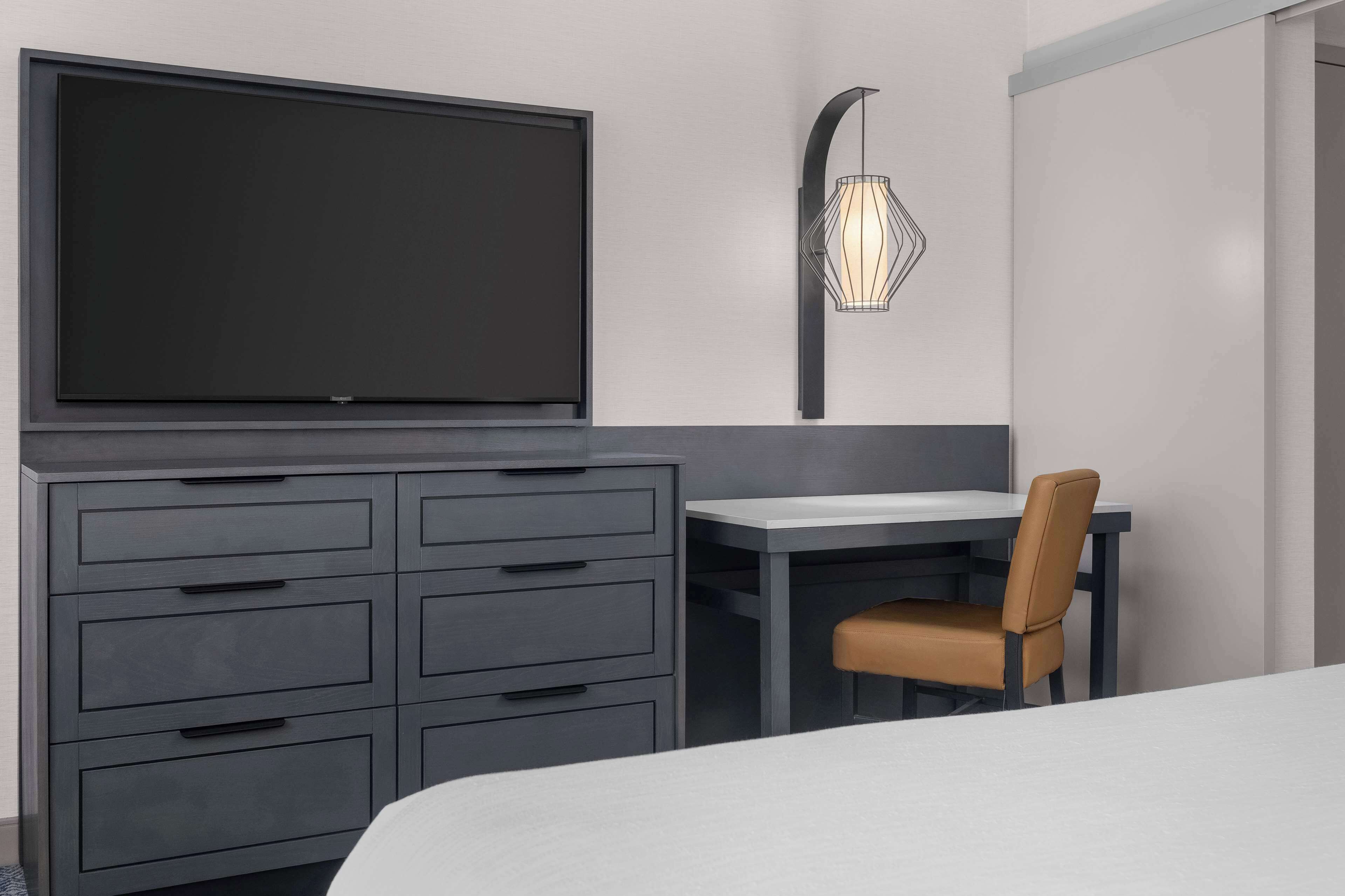 A modern hotel room with a large flat-screen TV mounted on the wall, a desk with a chair, and a decorative wall light.