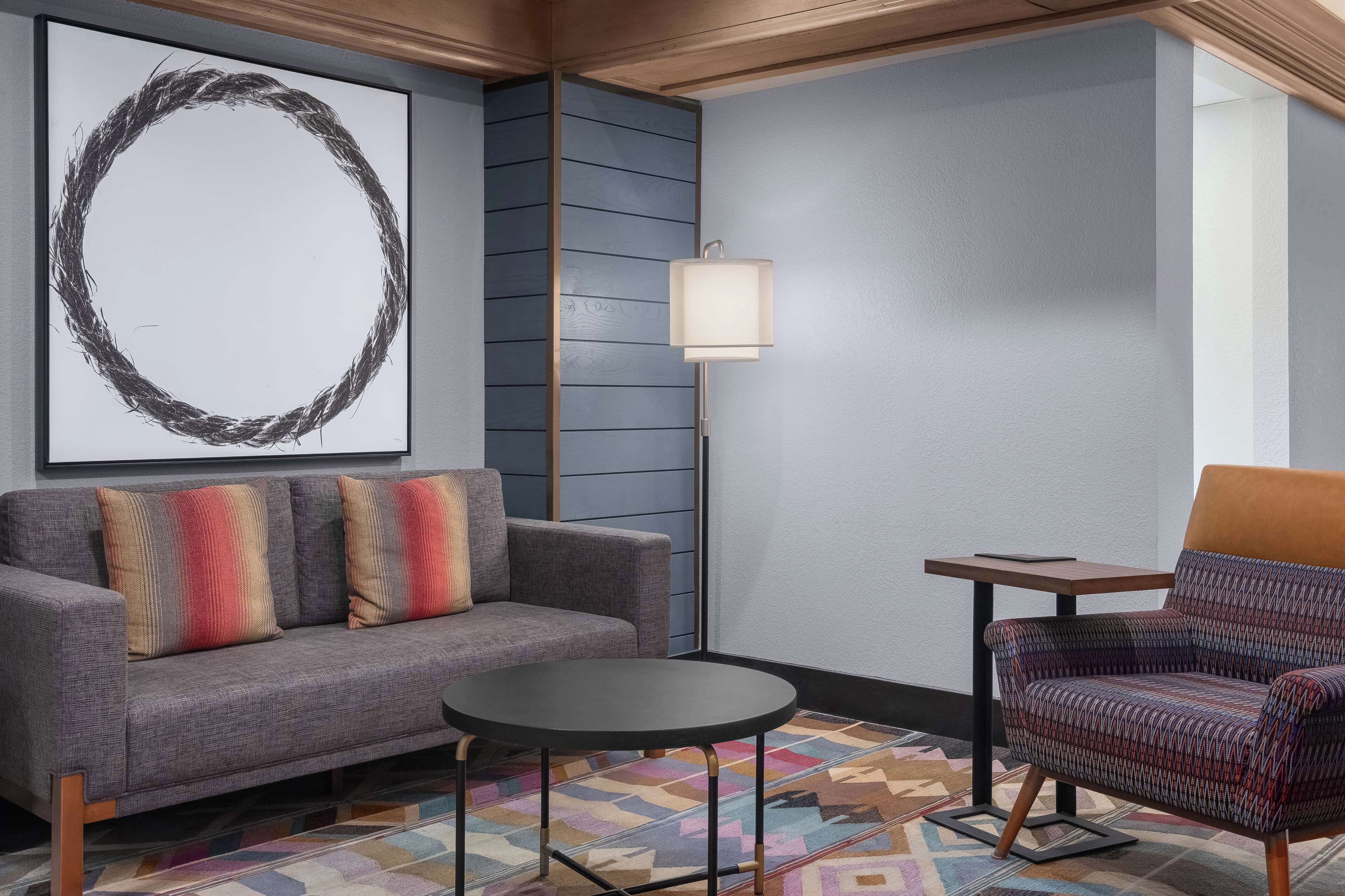 A modern living room with a gray sofa, colorful pillows, a patterned armchair, a small round table, and a floor lamp.