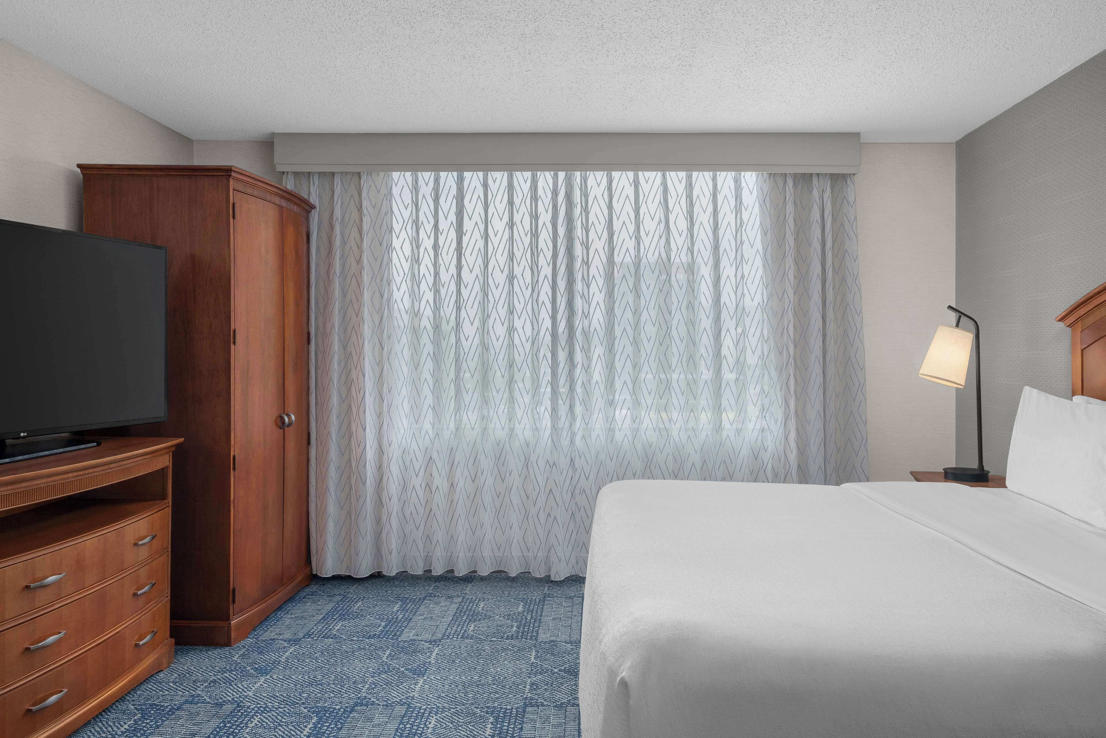 A hotel room with a large bed, a wooden wardrobe, a television on a stand, and a window with patterned curtains.