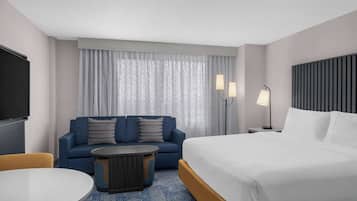 A hotel room with a bed, a sofa, a round coffee table, a TV, and two bedside lamps.