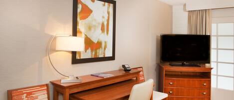 A hotel room with a wooden desk, a chair, a television, a lamp, and a framed abstract painting on the wall.