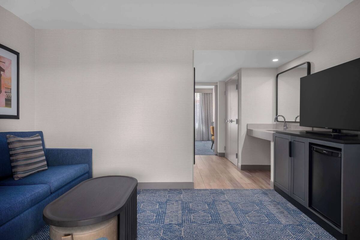 Uma sala de estar moderna com um sofá azul, uma mesa de centro e uma televisão. Um corredor leva a uma área de cozinha com uma pia e armários.