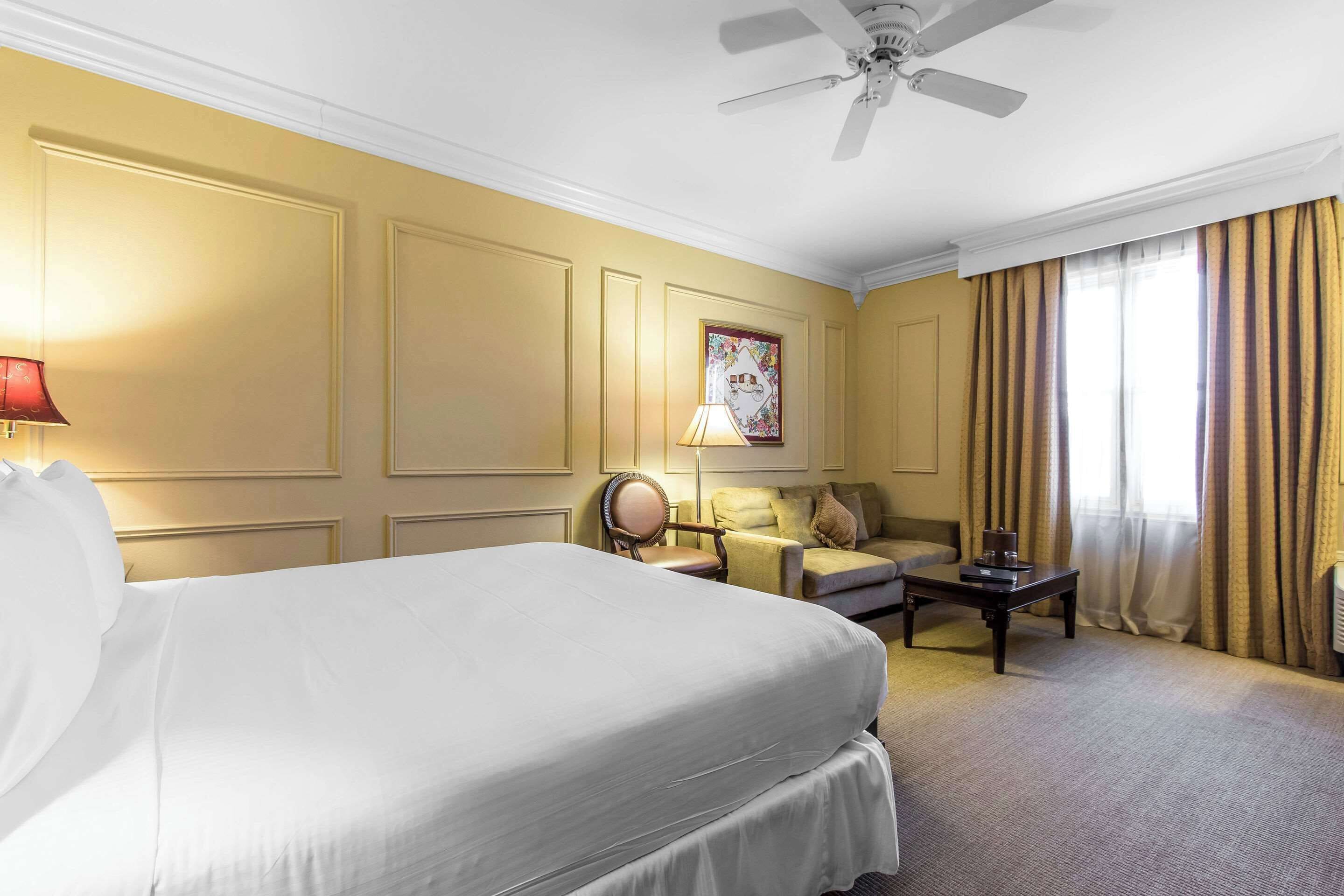 A hotel room with a large bed, a desk, a chair, a lamp, and a ceiling fan.