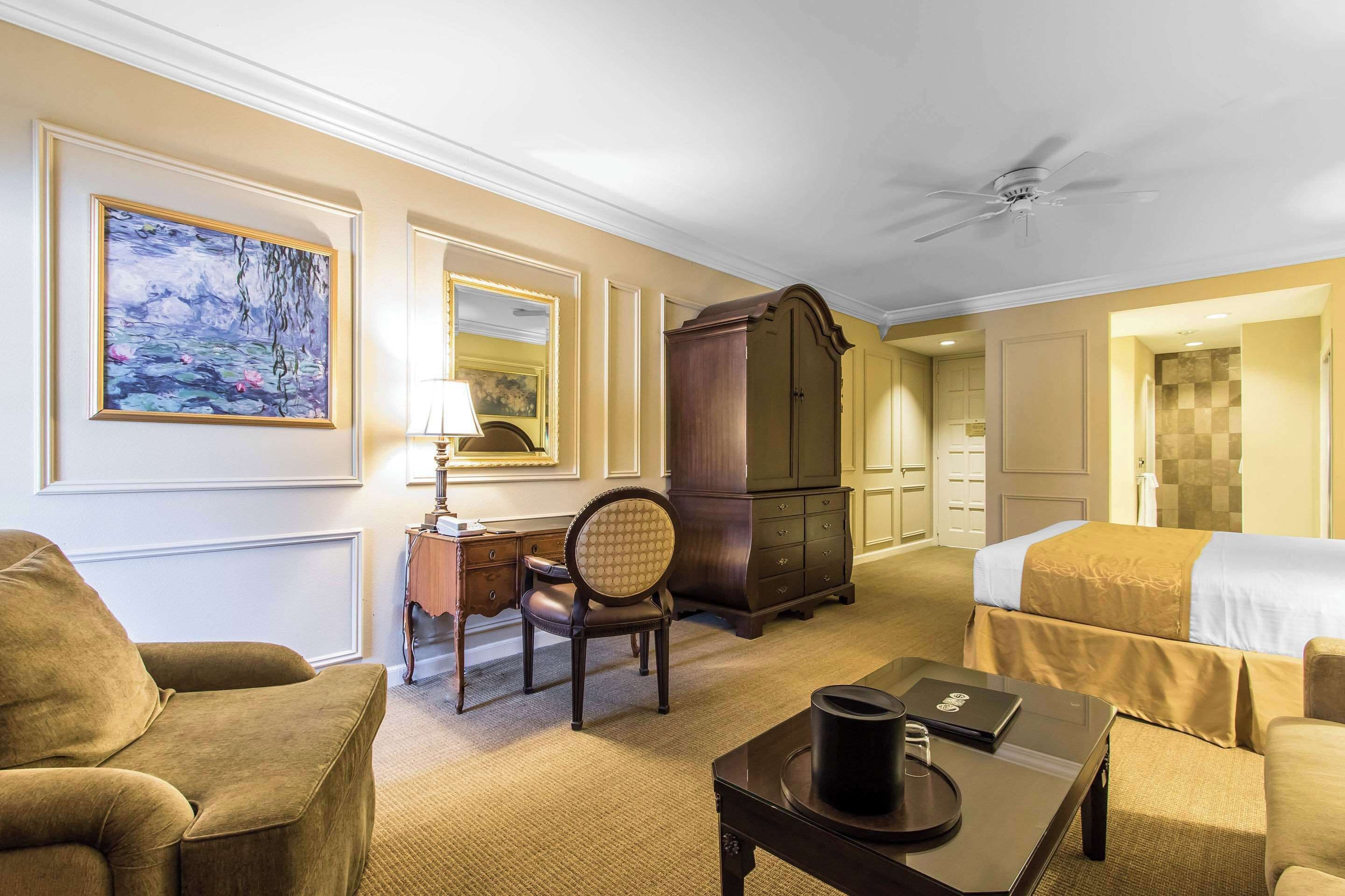 A hotel room with a bed, a desk with a lamp, a chair, a wardrobe, and a painting on the wall.