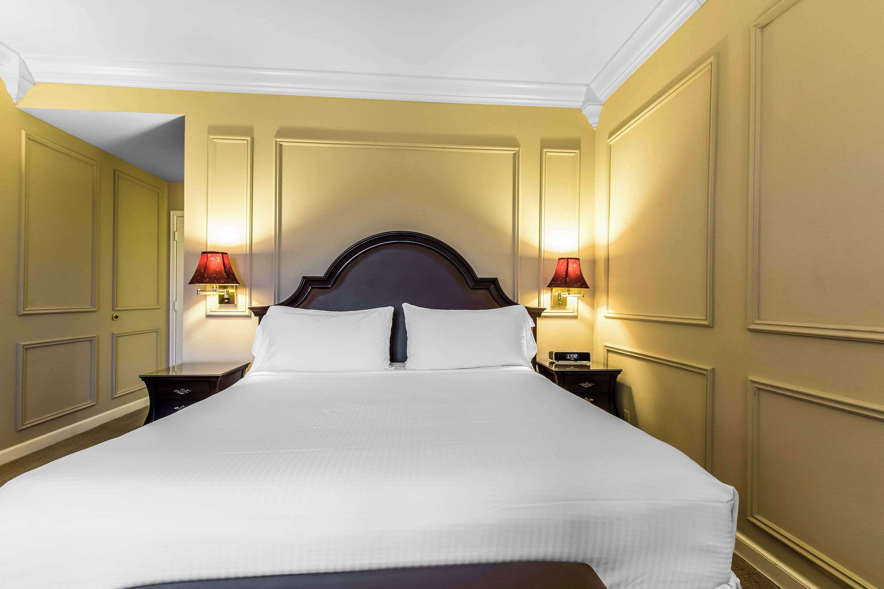 A hotel room with a large bed, two bedside tables with lamps, and cream-colored walls with paneling.