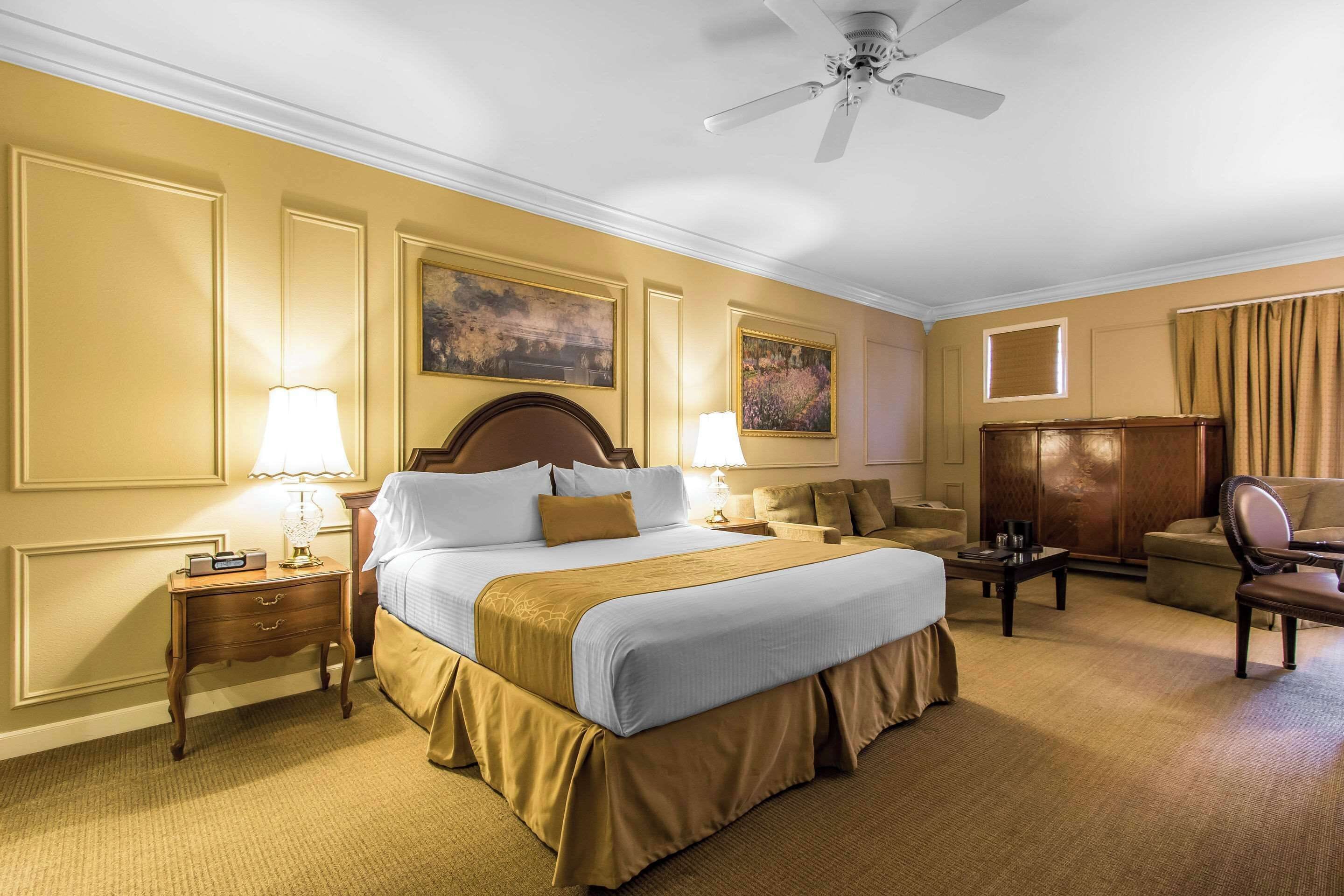 Habitación de hotel con una cama grande, dos mesitas de noche con lámparas, un armario de madera, una silla y un ventilador de techo.