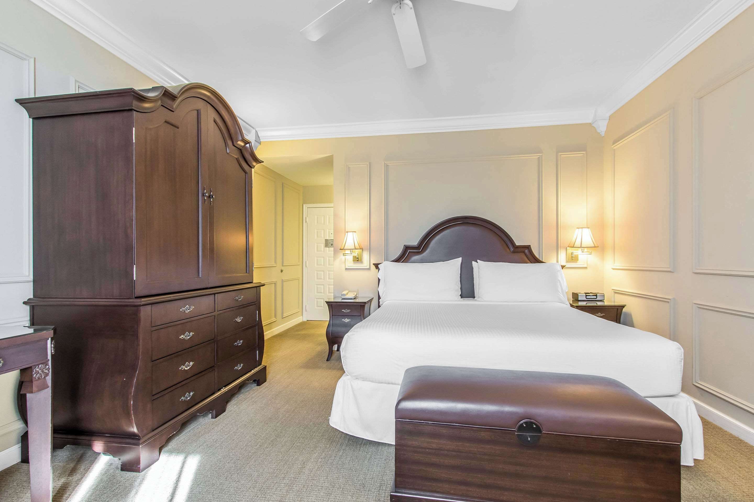 A hotel room with a large wooden dresser, a bed with white linens, a nightstand with a lamp, and a ceiling fan.