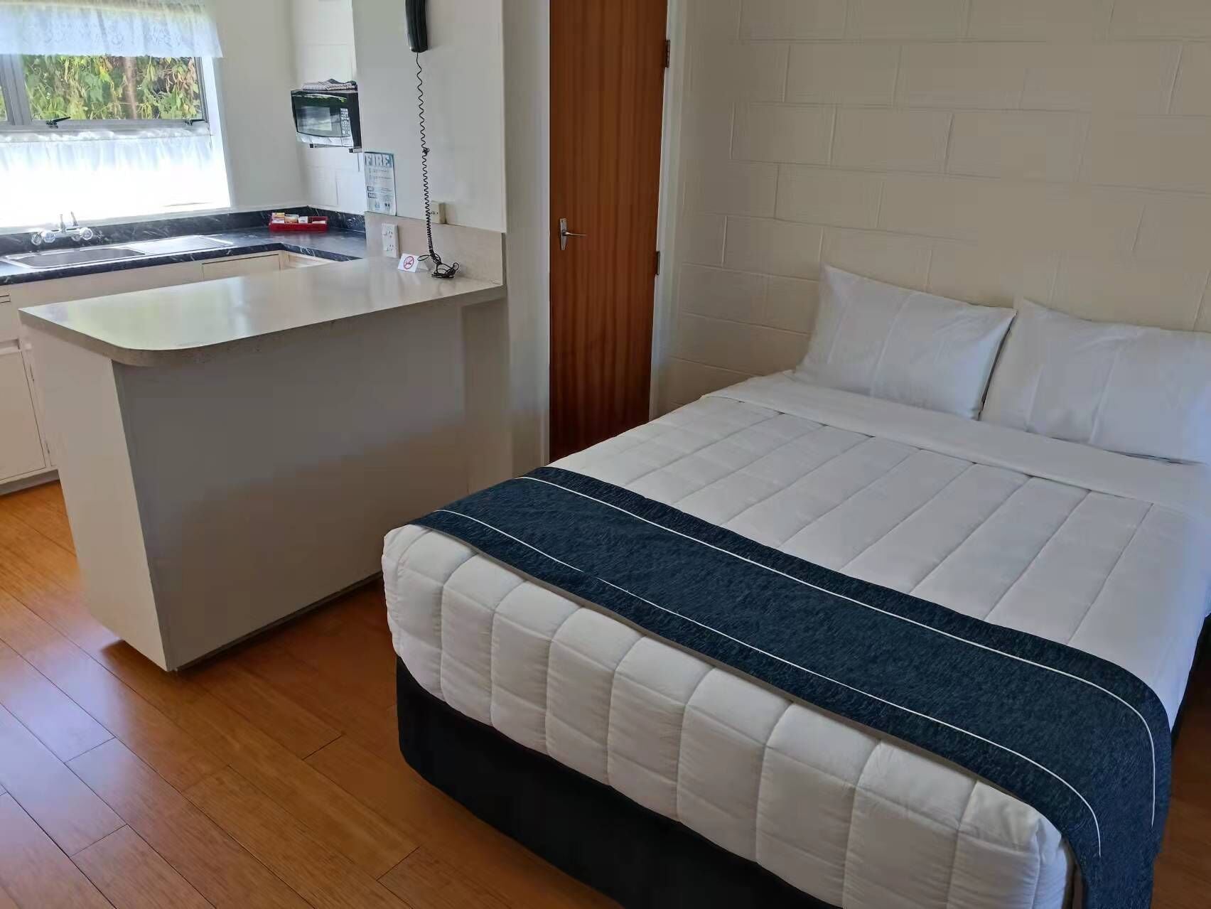 A bedroom with a bed, a bedside table, a small kitchenette with a sink, and a window.