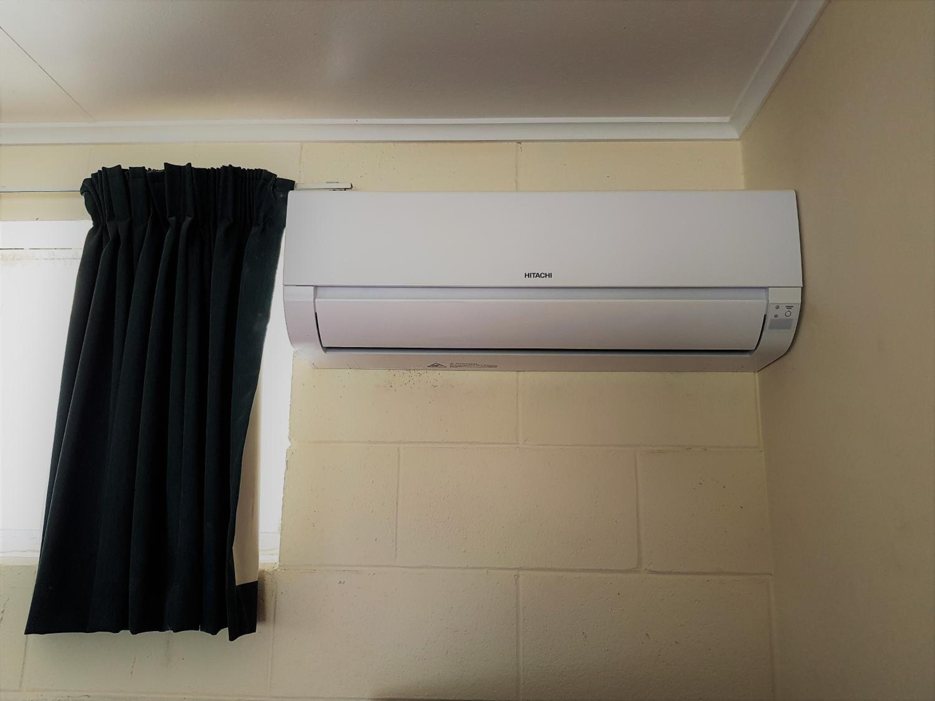 A room with a white air conditioning unit mounted on a beige wall, a black curtain, and a window with blinds.