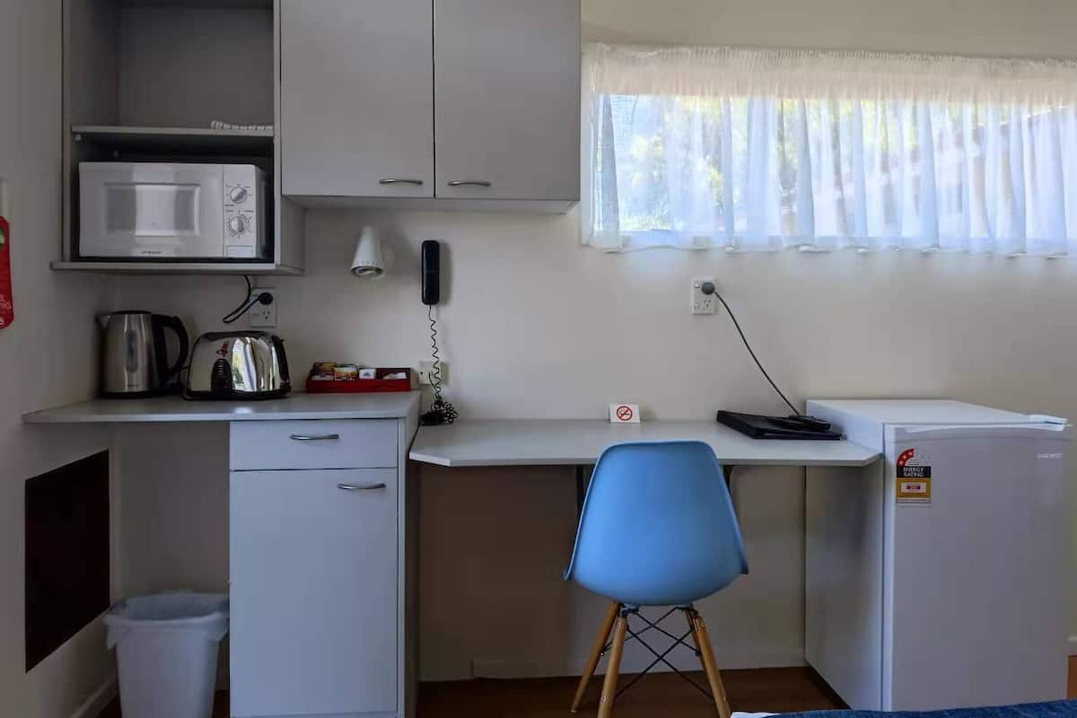 Una kitchenette compacta con microondas, hervidor de agua, tostadora y un refrigerador pequeño. Hay un escritorio con una silla, una ventana con cortinas translúcidas y un teléfono en la pared.