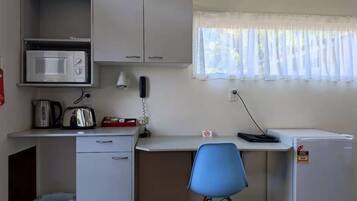 Una kitchenette compacta con microondas, hervidor de agua, tostadora y un refrigerador pequeño. Hay un escritorio con una silla, una ventana con cortinas translúcidas y un teléfono en la pared.