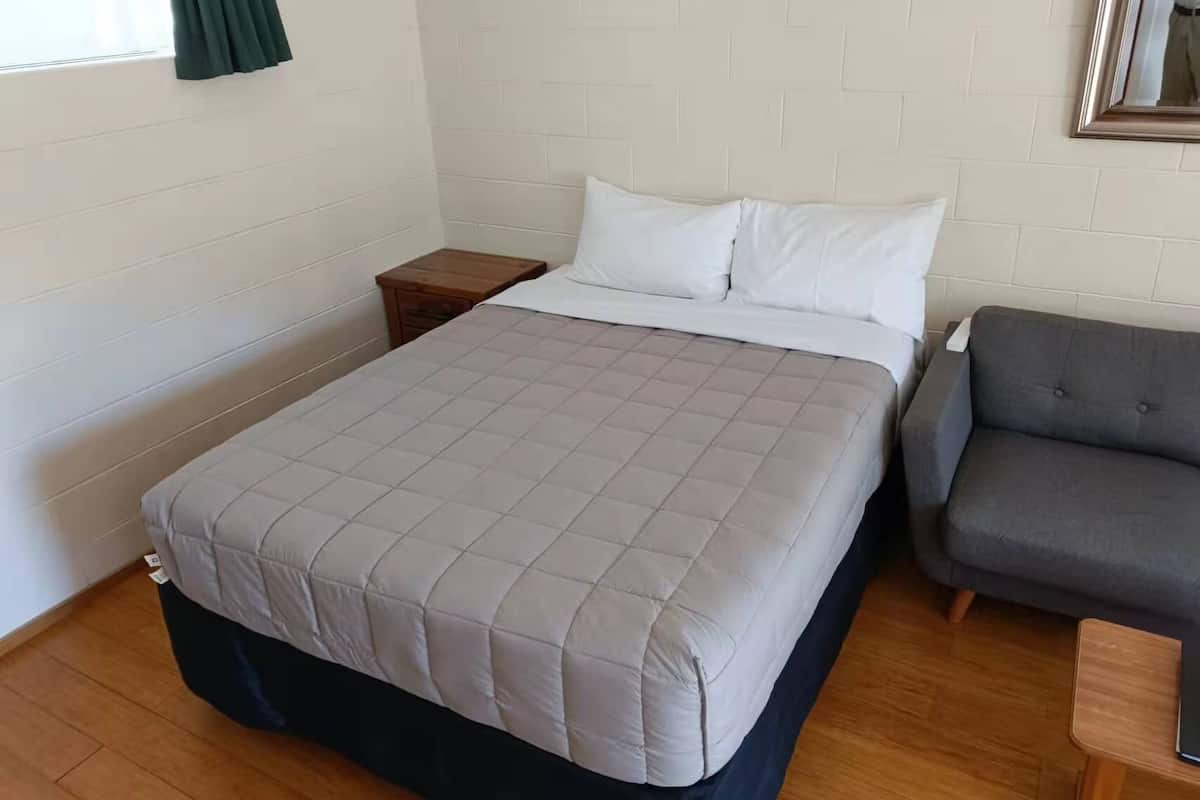 Una cama individual con un edredón gris, una mesita de noche de madera, un sofá gris y un espejo en la pared.