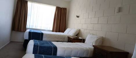 A hotel room with two beds, a brick-patterned headboard, a window with curtains, and a small bedside table.