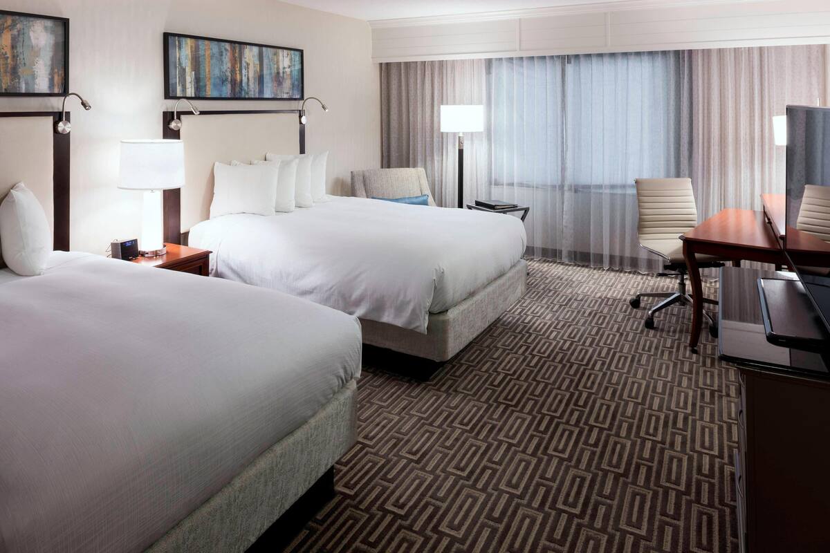 A hotel room with two beds, a desk, a chair, and a large window with curtains.
