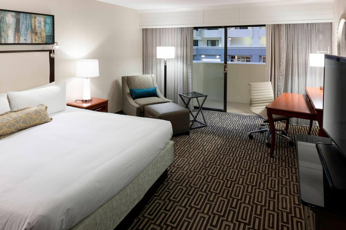 A hotel room with a large bed, two armchairs, a small table, a TV, and a balcony with curtains.