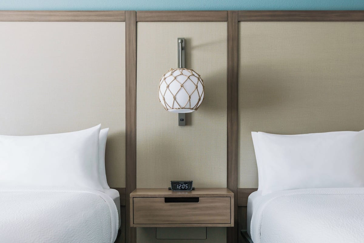 Habitación de hotel con dos camas, una mesita de noche con un reloj digital y una lámpara de pared.
