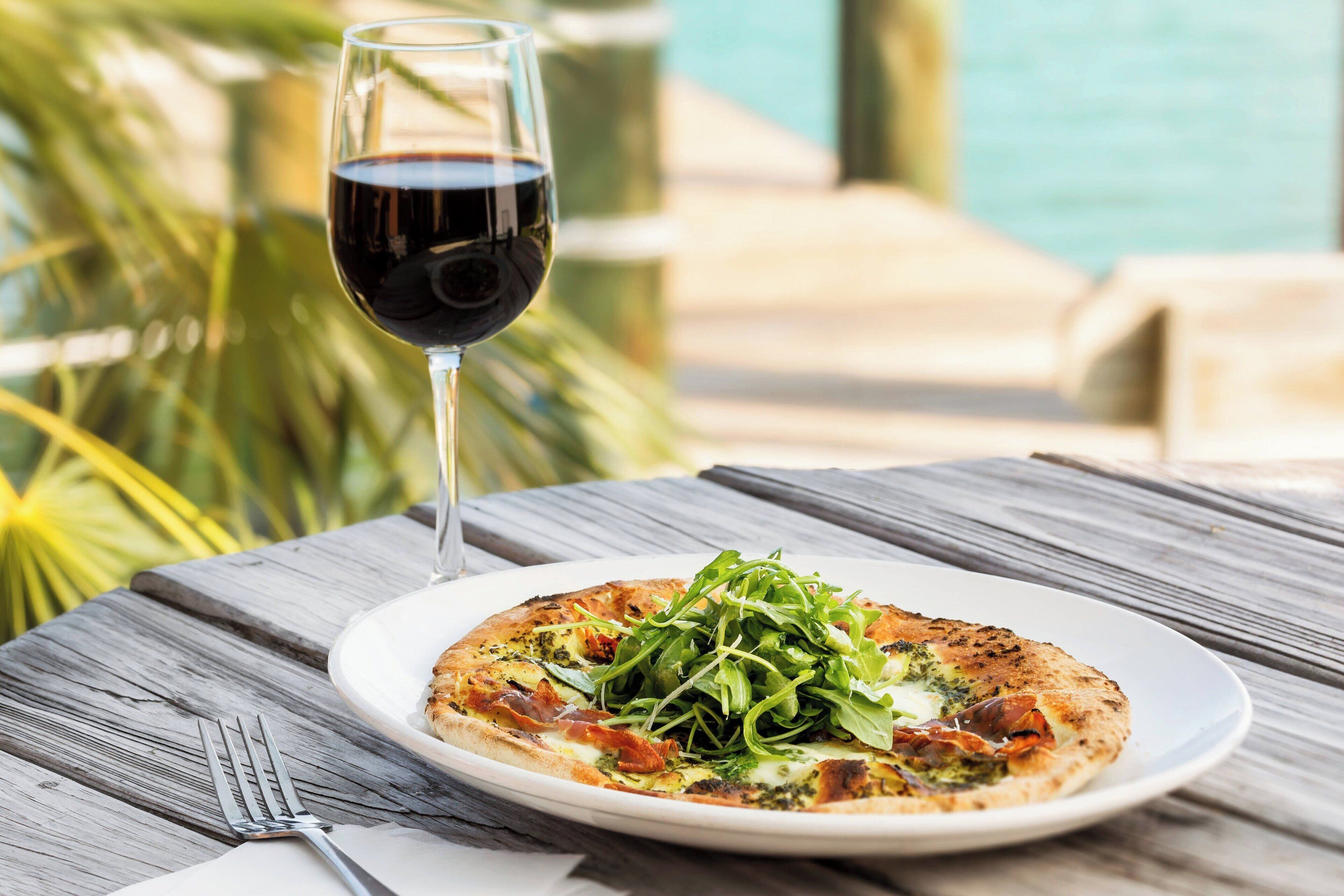 A plate of pizza with greens and cheese on a wooden table with a glass of red wine.