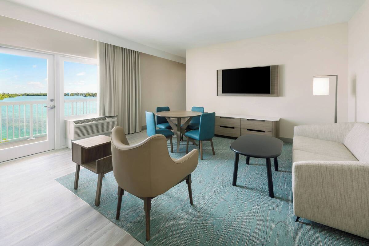 Habitación de hotel con balcón, zona de comedor, sofá, televisión y televisor de pantalla plana montado en la pared.