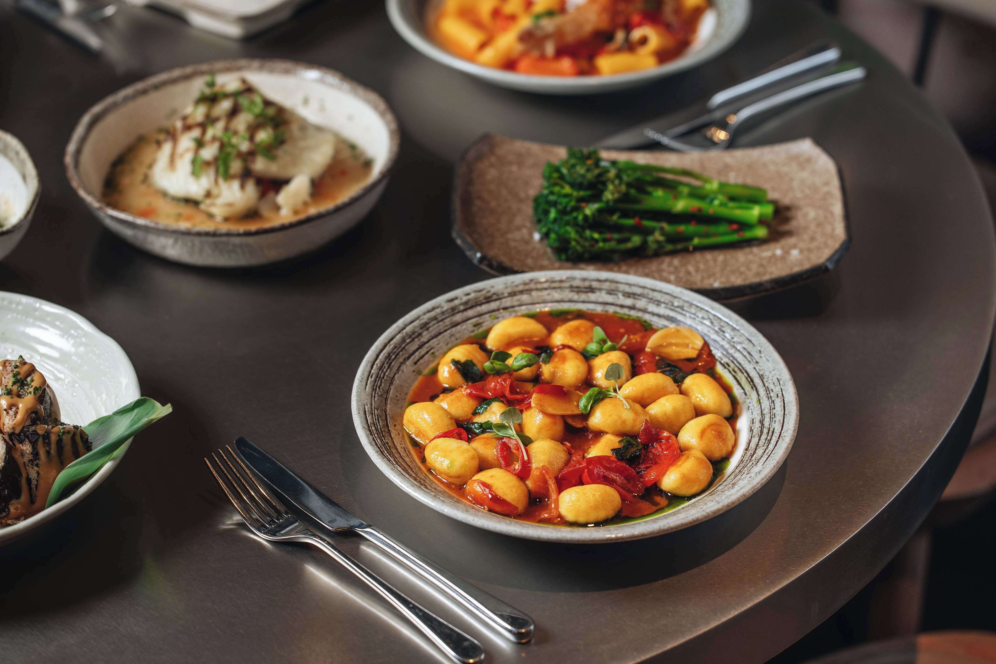 A table with various dishes, including a bowl of gnocchi with vegetables and a plate of fish.