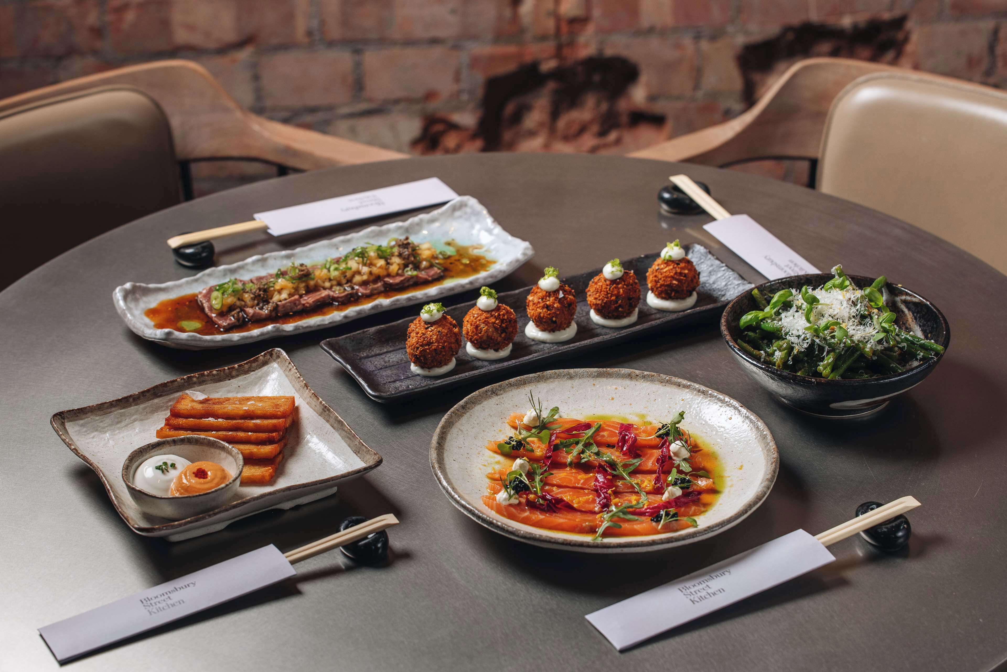 Una mesa dispuesta con diversos platos, incluyendo un bol de vegetales, un plato de carne y varios aperitivos pequeños y redondos.